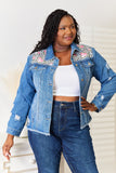 Woman wearing a denim jacket with floral embellishments and jeans, standing against a white background.