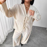Person wearing a cream-colored outfit with a tied front design in a minimal indoor setting.