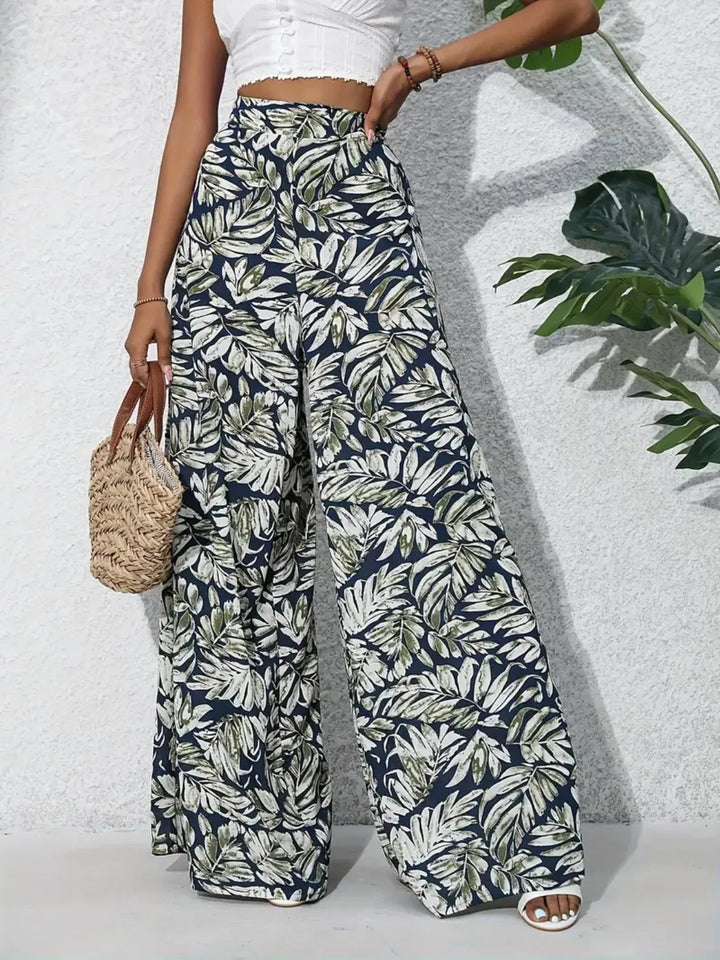 Person wearing tropical leaf print pants with a white top and sandals, holding a woven bag.