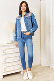 Woman wearing a blue denim jacket with floral embroidery and matching jeans in a room.