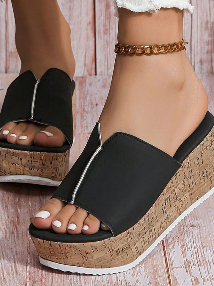 Black wedge sandals with cork platforms worn by a person on a wooden floor.