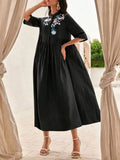 Woman wearing a black dress with floral embroidery standing in front of beige curtains.
