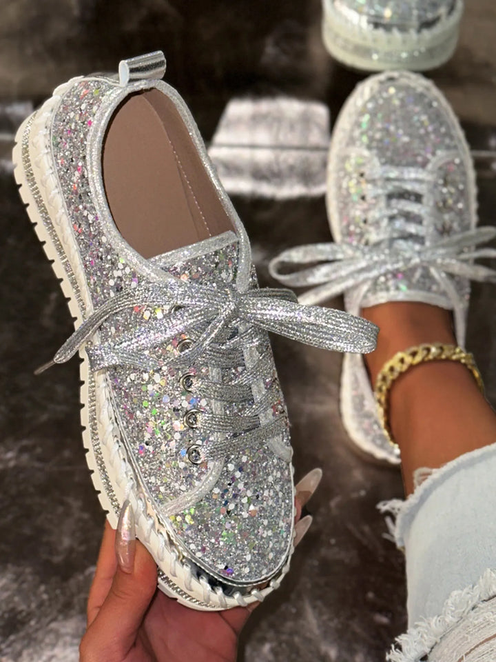 Silver glittery sneakers with rhinestone laces held by a hand against a blurred background.