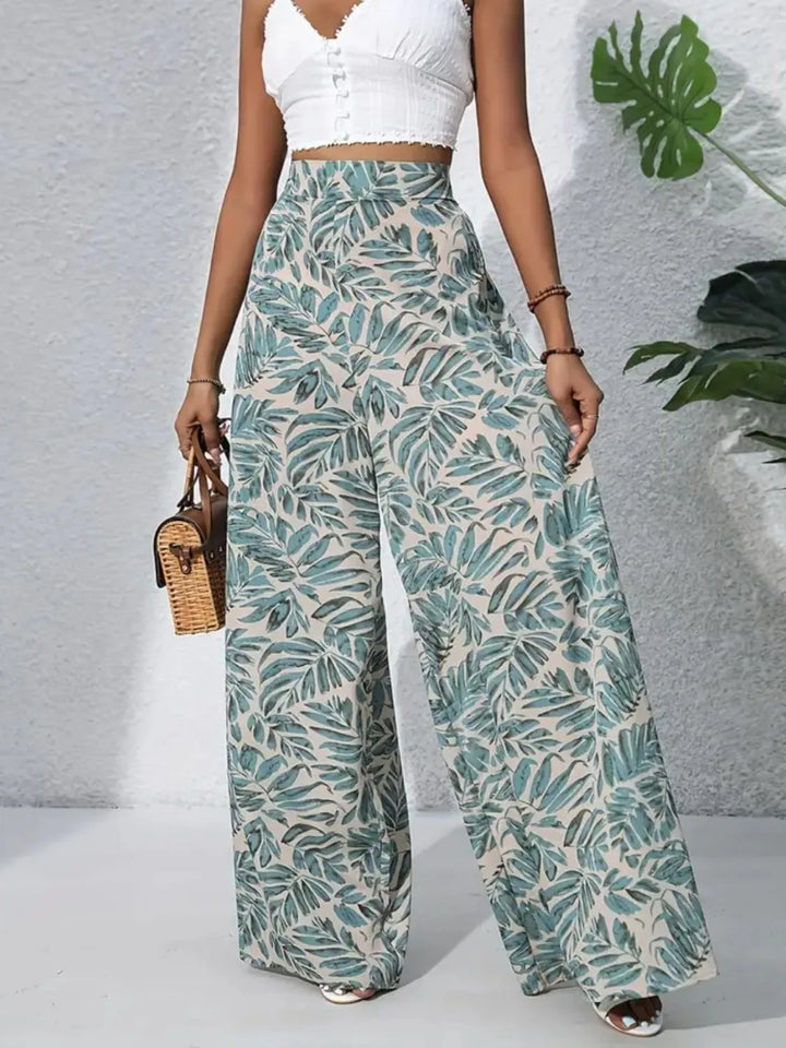 Person wearing leaf-patterned pants with a white top and small bag, standing against a light gray background.