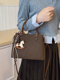 Brown handbag with a rocking horse charm held by a person wearing a blue sweater and plaid shirt.