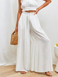 Person wearing a white outfit with wide-leg pants and a woven handbag indoors.
