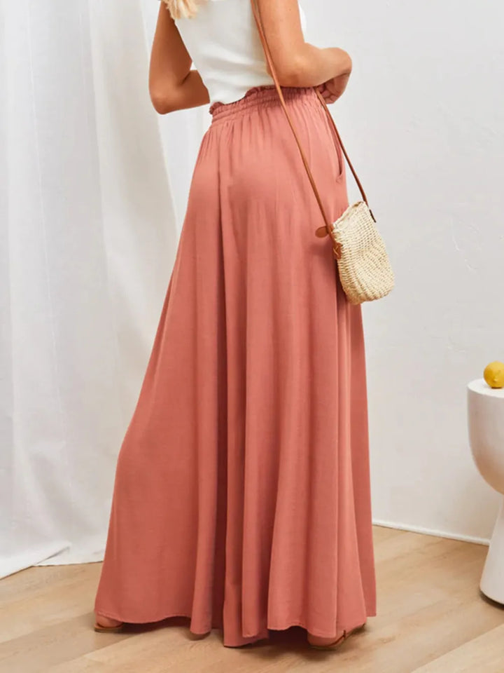 Person wearing a long pink skirt with a white top and carrying a small bag indoors.