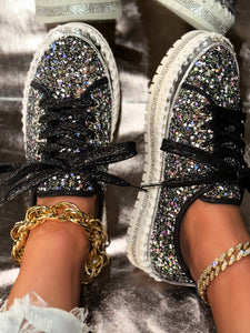 Silver glittery sneakers with rhinestone laces held by a hand against a blurred background.